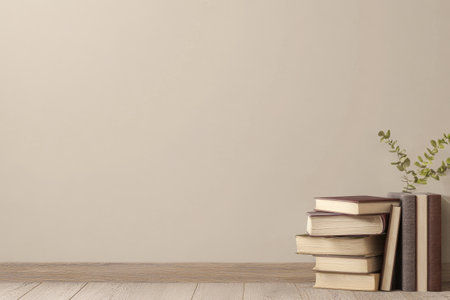 A collection of books neatly stacked on a wooden floor accompanied by a small green plant. The backdrop features a plain wall enhancing the serene atmosphere.の素材