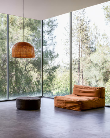 A cozy modern living space features a large window that brings in abundant natural light. The area includes a simple brown sofa and a round ottoman surrounded by peaceful greenery.の素材