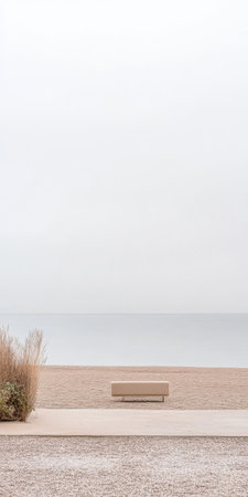 A serene beach scene features a minimalist bench on a sandy shore surrounded by gentle grasses. The water meets the horizon under a soft cloud-covered sky creating a peaceful atmosphere.の素材