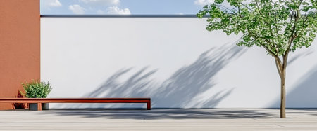 Bright outdoor area features a clean white wall a wooden bench and a lush green tree. Shadows dance on the ground under clear skies during the midday light creating a tranquil atmosphere.の素材