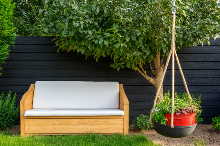 A wooden bench sits comfortably beneath a thriving tree complemented by a red hanging planter with vibrant plants. The setting showcases a peaceful garden atmosphere.の素材