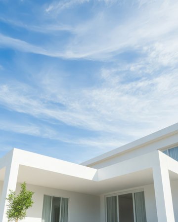 A white modern house appears against a bright blue sky filled with wispy clouds. Sunlight highlights the sharp lines and minimalist design of the structure creating a tranquil atmosphere.の素材