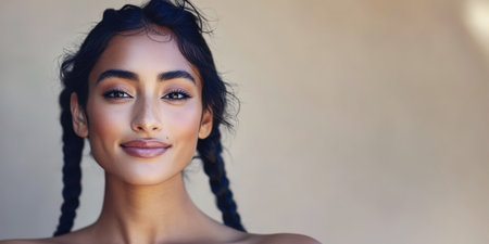 A young woman with long dark hair in braids smiles warmly while standing in front of a neutral-colored wall. Soft natural light highlights her features creating a friendly and inviting atmosphere.の素材