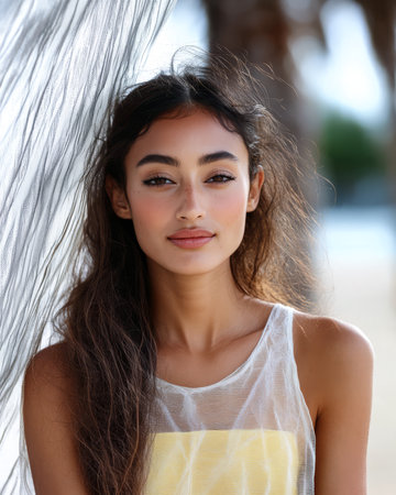 The young woman with flowing hair stands confidently against a sheer backdrop with a beach view behind her. The sun casts a warm glow enhancing the tranquil setting.の素材