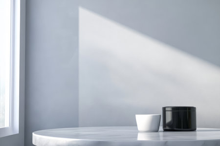 Sunlight filters through a window casting a soft shadow on a light gray wall. A marble table holds a small white cup next to a sleek black container creating a serene atmosphere.の素材