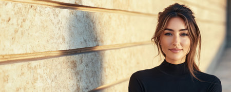 A young woman stands with a relaxed expression against a beautiful stone wall. Soft golden hour light enhances her features creating a warm and inviting atmosphere.の素材