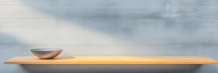 A light wooden shelf holds a smooth gray bowl bathed in soft natural light. The background features a subtle textured wall creating a serene atmosphere.の素材