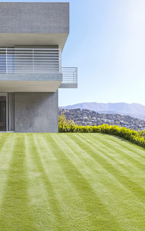 A sleek modern house features a well-manicured lawn with striped grass patterns. The property has expansive views of surrounding hills under clear blue skies.の素材