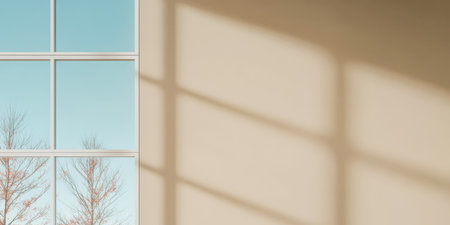 Sunlight streams through a large window casting soft shadows on a light-colored wall. Outside trees with subtle foliage create a serene atmosphere on a clear bright day.の素材