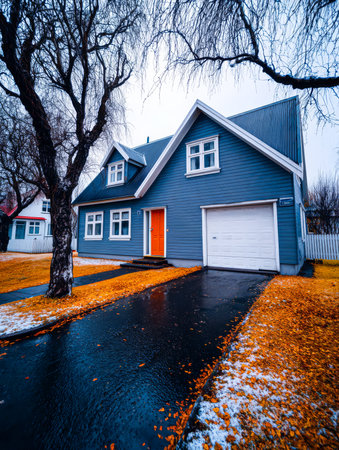 A charming blue house features an orange door and sits in a serene neighborhood. Autumn leaves are scattered on the ground highlighting the beauty of the season against a grey sky.の素材