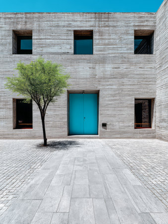A minimalist building with grey concrete walls features a striking blue door at the center. The surrounding area showcases a small tree and stone paving all under clear blue skies.の素材