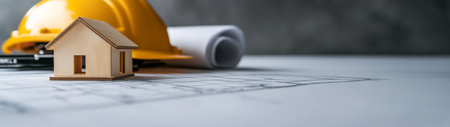 A small wooden model house is placed on a drafting table alongside a rolled-up blueprint and a hard hat. The workspace is designed for construction planning and design specifications.の素材
