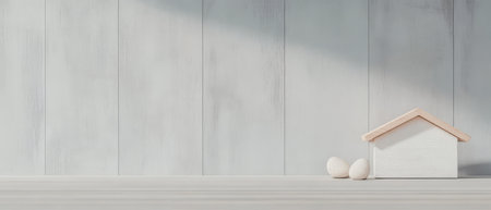 A small white house model with a wooden roof sits alongside two decorative eggs on a smooth wooden surface. Soft natural light illuminates the scene creating a tranquil atmosphere.の素材