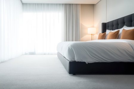 A spacious and minimalist bedroom features a comfortable bed with plush pillows illuminated by soft natural light streaming through sheer curtains. The clean design creates a serene atmosphere.の素材