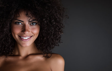 A cheerful woman with voluminous curly hair displays a warm smile while posing against a dark backdrop. Her confident expression highlights her natural beauty and personality.の素材