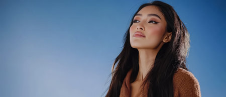 A woman with long dark hair stands confidently while looking up basking in the sunlight. The blue sky creates a serene backdrop capturing a moment of tranquility and poise.の素材