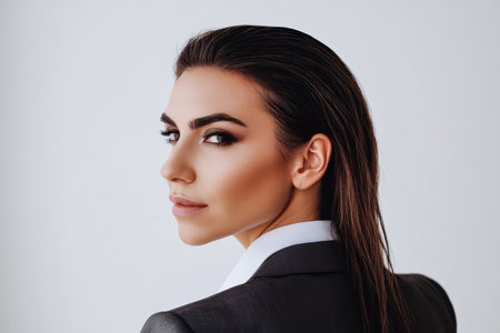 A woman with sleek hair and elegant makeup is dressed in a tailored suit, glancing back with a confident expression. The neutral background enhances her professional appearance.の素材