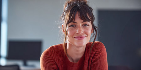 A young woman with dark hair smiles warmly while seated at a desk in a contemporary office space. The atmosphere is bright and inviting, ideal for collaboration and creativity.の素材