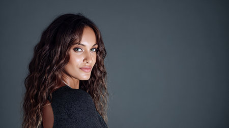 A woman with long, wavy hair looks over her shoulder with a confident smile. The neutral background enhances her striking features and showcases her stylish outfit.の素材