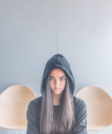 A girl with long hair wears a black hoodie and looks serious while sitting in a minimalist setting featuring wooden chairs and soft ambient light. The atmosphere hints at introspection.の素材