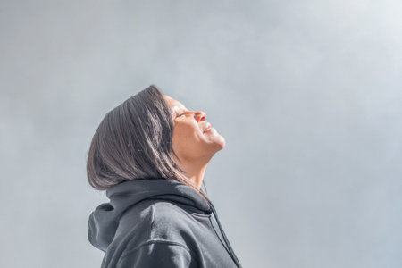 A woman stands outdoors, tilting her head back and smiling as sunlight illuminates her face. She wears a casual black hoodie and seems at peace in the warm atmosphere.の素材