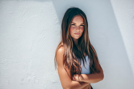 A young woman stands confidently with her arms crossed against a white wall. Her long hair and freckles highlight her natural charm under bright sunlight.の素材