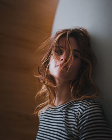 A woman with wavy hair stands against a lightly curved wall, exuding a contemplative vibe. The warm tones of the background create a serene atmosphere, emphasizing her thoughtful expression.の素材