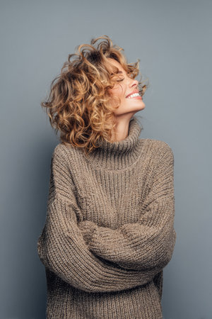 A woman with curly hair smiles joyfully while crossing her arms. She wears a chunky, knitted sweater in a neutral tone, captured against a plain backdrop that enhances her cheerful demeanor.の素材