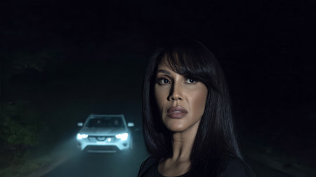 A woman with long dark hair looks intently at an approaching vehicle on a dimly lit road. The scene conveys a feeling of suspense and mystery under the night sky.の素材
