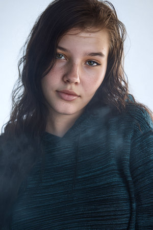 A young woman with long, wavy hair is captured in a relaxed pose, wearing a cozy hoodie while soft smoke envelops the background, creating an intriguing atmosphere.の素材
