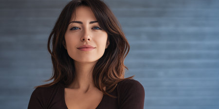 A young woman with long brown hair stands confidently in front of a textured blue wall. She has a calm expression and a gentle smile, dressed in a simple top. The atmosphere is serene.の素材