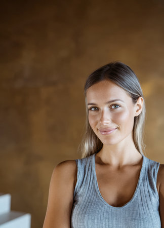 A young woman with long hair smiles confidently while wearing a casual gray tank top. The warm, simple background enhances her cheerful demeanor and bright expression.の素材