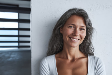 A woman with gray hair smiles warmly while standing against a white wall in a contemporary interior. Sunlight fills the room, creating a bright atmosphere.の素材