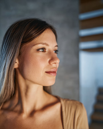 A young woman stands quietly, looking to the side with a serene expression. Soft light highlights her features in a contemporary setting with wooden elements.の素材