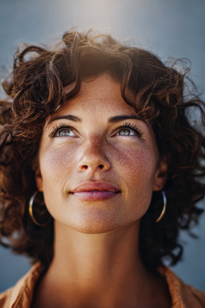 A woman with curly hair and freckles looks upward with a serene expression, basking in the sunlight in a calm outdoor environment. She appears contemplative and relaxed.の素材