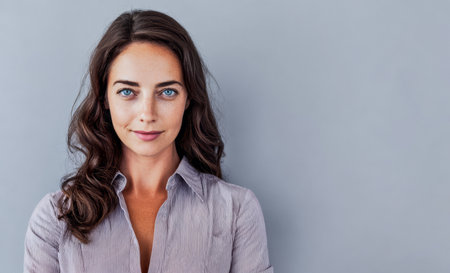A woman with long dark hair stands confidently against a smooth gray wall. She wears a light shirt and has a friendly expression, looking directly at the viewer with bright blue eyes.の素材