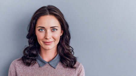 A woman with dark, wavy hair smiles warmly while wearing a light sweater and a collared shirt. She stands against a soft gray wall, creating a friendly atmosphere.の素材