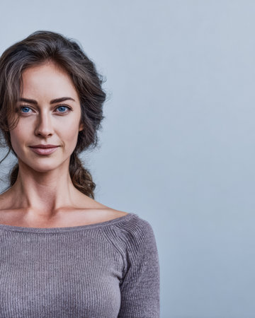 A woman with long, flowing hair and striking blue eyes smiles softly while wearing a fitted sweater. She stands against a soft blue wall, showcasing her natural beauty.の素材