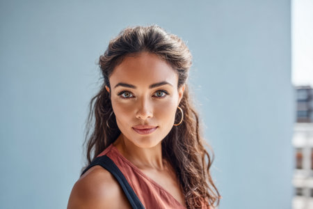 A woman with curly hair stands facing the camera, looking confidently. She wears a casual top and hoop earrings, with a soft smile. The bright blue background adds to the cheerful atmosphere.の素材