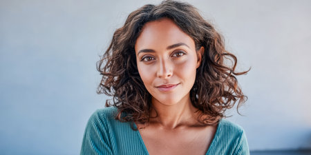 A young woman with curly hair stands smiling softly while wearing a blue top. She is in a well-lit area with a plain background that enhances her natural beauty.の素材