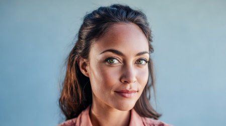 A woman stands confidently, showcasing her long dark hair and vibrant green eyes. She sports a soft pink shirt and poses against a textured light blue background, creating a calm atmosphere.の素材