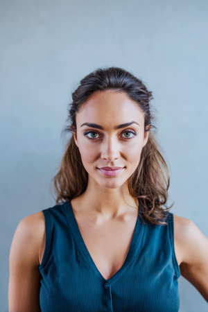 A woman with long, flowing hair and a confident smile poses against a soft gray wall. She wears a dark sleeveless top, exuding a sense of poise and elegance.の素材