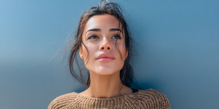 A young woman with dark hair and clear skin looks up with a serene expression. She is wearing a cozy sweater and standing in front of a solid blue background on a sunny day.の素材