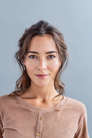 A young woman with long brown hair smiles softly while wearing a simple brown top. Her natural makeup enhances her features, set against a plain gray background.の素材