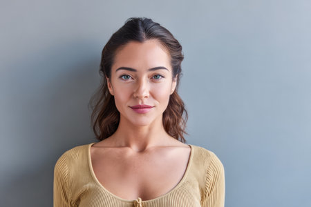 A young woman stands confidently in front of a grey wall, showcasing her natural beauty and calm expression. Her casual outfit highlights her relaxed demeanor in the soft morning light.の素材