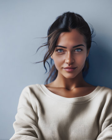A young woman with long brown hair and bright blue eyes sits calmly against a soft gray wall, wearing a cozy light sweater. She exudes confidence and warmth.の素材