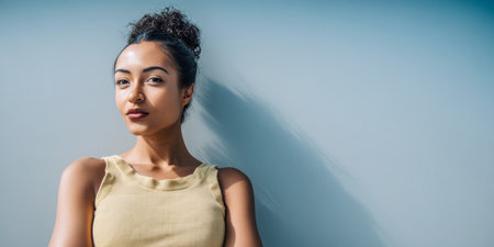 A woman with curly hair stands confidently against a blue wall, wearing a light-colored top. The sunlight highlights her features, creating a vibrant atmosphere.の素材