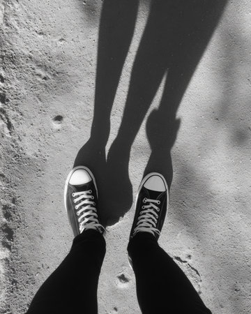 Black sneakers with white laces stand on a sandy surface, casting elongated shadows in the sunlight. The scene captures a moment of leisure on a warm day.の素材