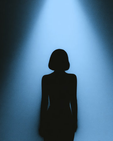 A silhouette of an individual with short hair stands in a dim environment. A beam of blue light shines down, creating a dramatic contrast against the dark background.の素材
