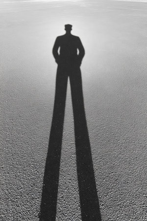 A tall shadow cast by a person standing with hands in pockets on a smooth surface. The bright sunlight creates a stark contrast between the shadow and the ground.の素材